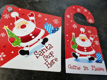 Load image into Gallery viewer, Two Christmas-themed door hangers with Santa Claus on a dark surface.