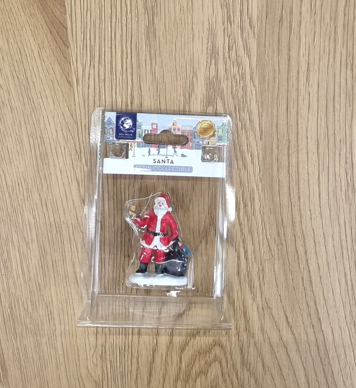 A Santa Claus miniature figurine in its clear plastic blister packaging, displayed on a light wood grain surface.