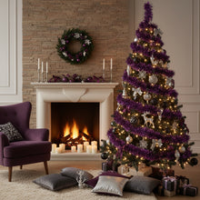 Load image into Gallery viewer, A decorated Christmas tree in a cozy living room beside a lit fireplace. The tree is wrapped in metallic purple tinsel with warm white string lights, silver and white ornaments, reindeer figures, and bows, with wrapped gifts underneath.