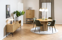 Load image into Gallery viewer, A modern dining room setup featuring a round wooden table with a ribbed pedestal base, surrounded by six dark green upholstered chairs on a round white rug. The room has light wood herringbone flooring, a slatted wood cabinet, and a multi-light pendant chandelier.