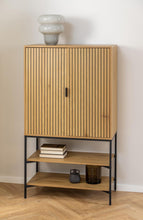 Load image into Gallery viewer, A modern wooden cabinet with a slatted double-door upper section, open lower shelves, and a black metal frame, shown against a plain wall on a wood floor with decorative objects on and around it.