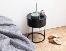 Load image into Gallery viewer, Black round side table with decorative items next to a bed with gray bedding on a wooden floor.