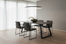 Load image into Gallery viewer, A modern dining room setup featuring a rectangular dark wood table with black metal legs, surrounded by four matching black upholstered chairs. The room has light wood flooring, a gray wall, sheer white curtains, and a circular black pendant light overhead.