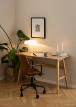 Load image into Gallery viewer, A warm, modern home office corner featuring a light wood desk with a brown leather swivel chair, styled with a lit table lamp, candles, and a framed print on the beige walls.