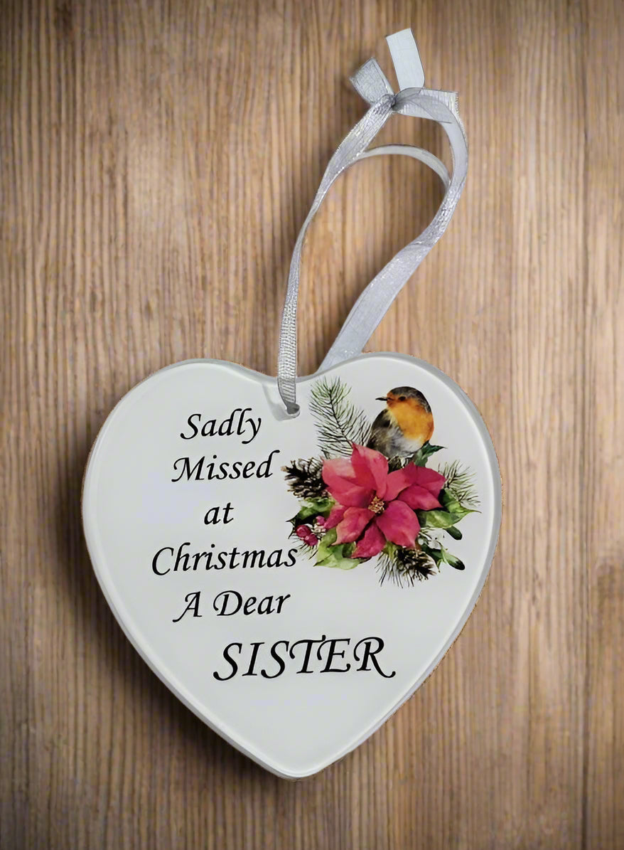 A heart-shaped white ceramic Christmas ornament hanging on a wooden surface, printed with the phrase 'Sadly Missed at Christmas A Dear SISTER' in black script, decorated with a robin bird, pink poinsettias, and green pine branches, and suspended by a white ribbon.