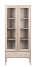 Load image into Gallery viewer, A tall, narrow light oak wood display cabinet with two glass doors, four interior shelves, and tapered legs, shown against a plain white background.