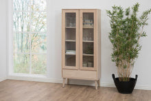 Load image into Gallery viewer, A tall light wood display cabinet with two glass-front upper doors and a lower drawer, shown in a bright room beside a window and a potted plant.
