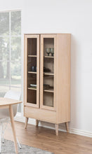Load image into Gallery viewer, A light natural wood cabinet with two glass-front upper doors and a lower drawer, shown in a bright room with a window, table, and wood floor.