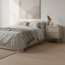 Load image into Gallery viewer, A minimalist bedroom scene with a neatly made bed in light beige linens and a gray throw blanket, beside a matching beige nightstand holding a small vase with dried grass and books. The room has pale wood flooring and a soft abstract wall art piece above the bed.