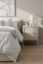 Load image into Gallery viewer, A modern bedroom scene featuring a light beige two-drawer nightstand beside a bed with white bedding, a white table lamp, and a small potted succulent, set against a warm beige wall with soft sunlight shadows.