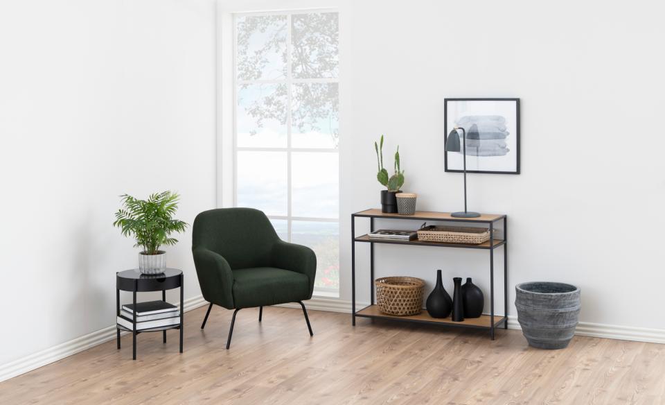 A styled interior room scene with a dark green armchair, a black side table with a plant, a narrow console shelf with decor, and a gray planter on a light wood floor against white walls.