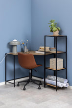 Load image into Gallery viewer, A modern L-shaped desk setup in a blue room corner, featuring a black metal frame with light wood desktops, a brown swivel office chair, a gray desk lamp, books, a plant, and a shelving unit holding storage boxes and rolled papers.