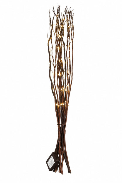 A decorative arrangement of tall, slender brown branches or twigs with warm white string lights woven through them, standing on a dark wooden surface against a plain off-white wall.