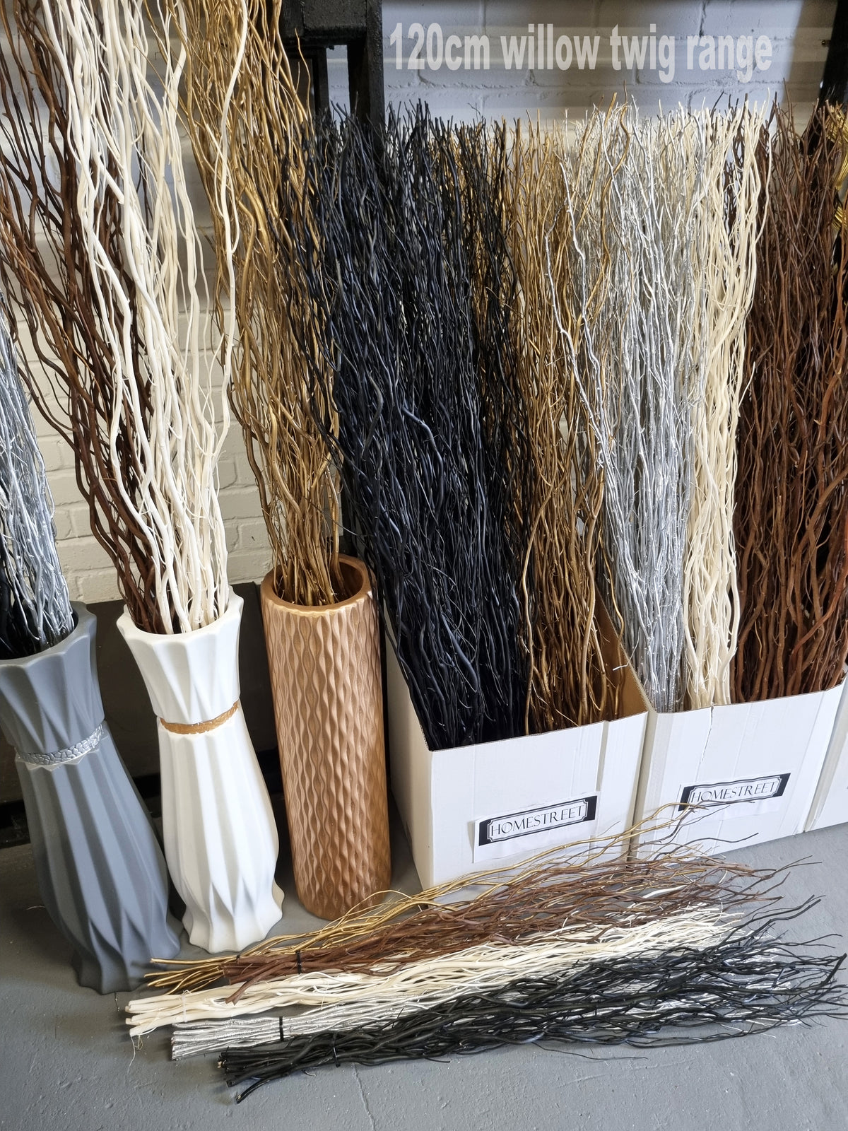A display of decorative dried willow twig arrangements in various colors and textures, shown in tall textured vases and white bins labeled 'SNOWSHIRE' and 'CANADA'. A bundle of loose brown and beige twigs lies on the floor in front.