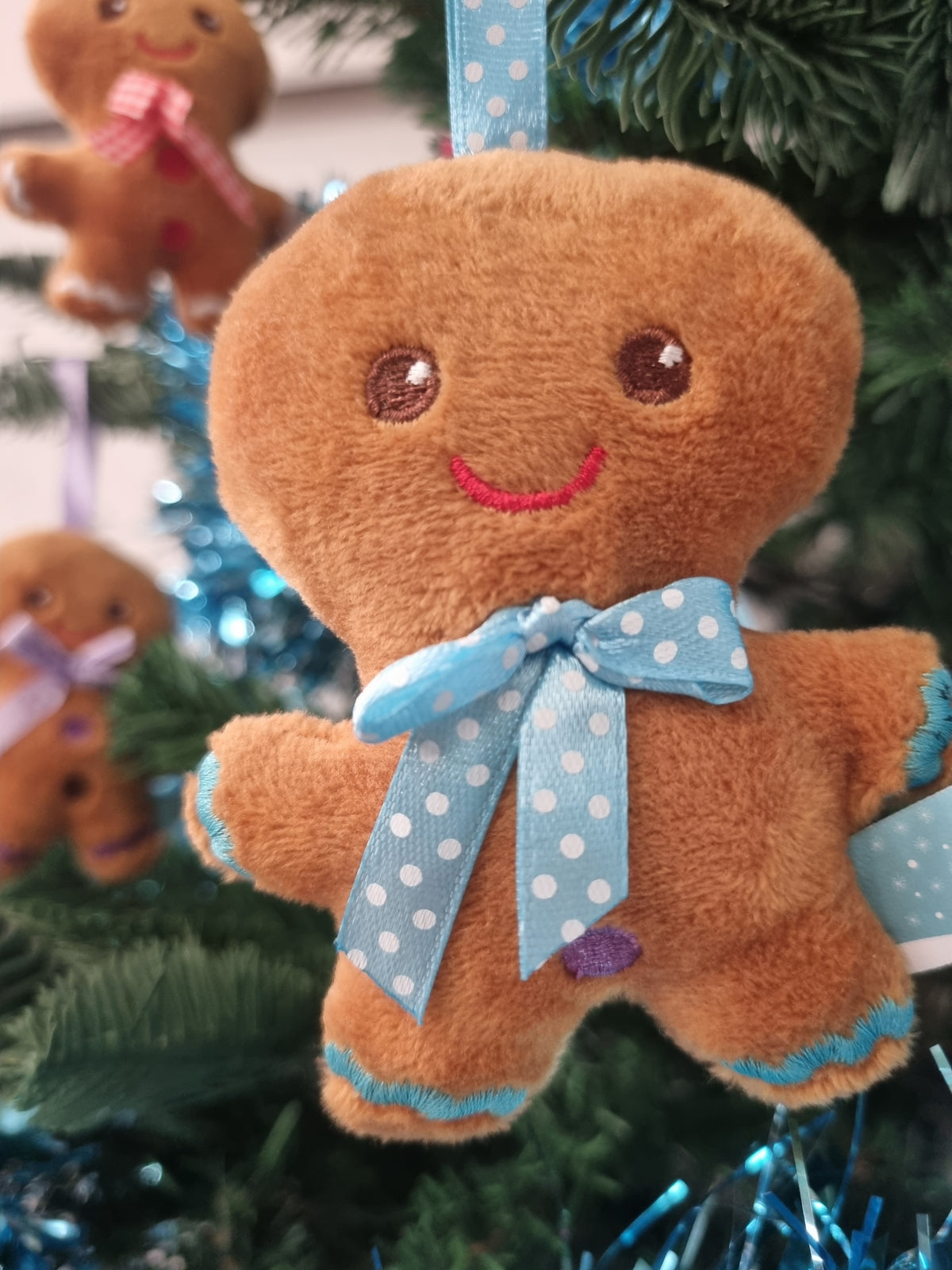 A brown plush gingerbread ornament hanging on a Christmas tree, with a light blue polka-dot bow around the neck and blue trim along the edges.