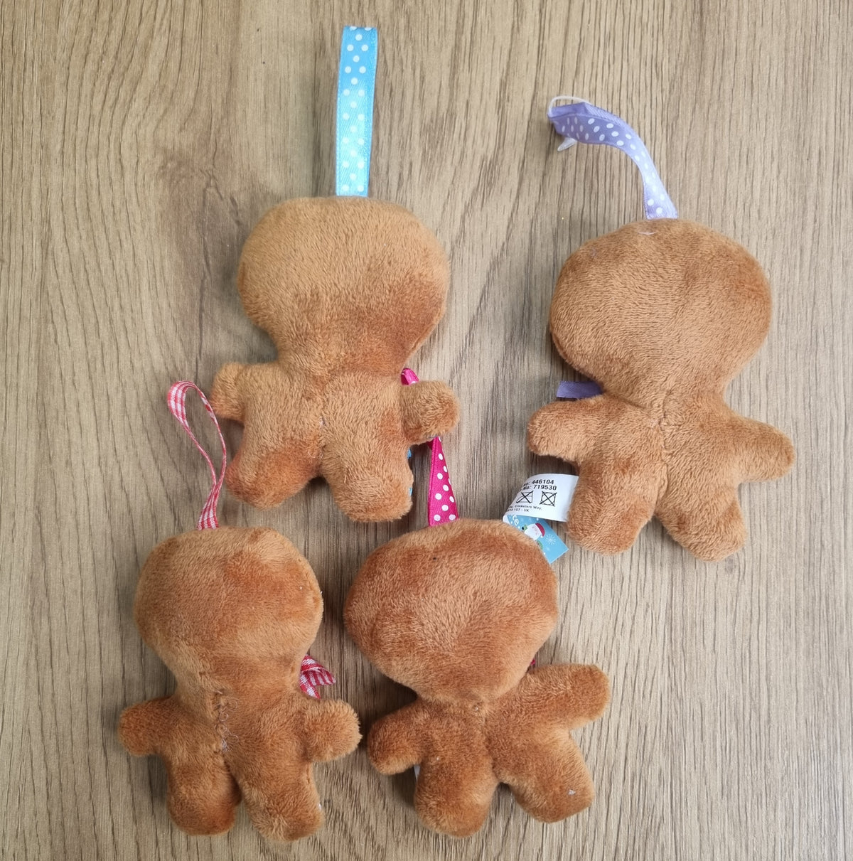 Four small plush gingerbread person ornaments or toys in brown fabric, arranged on a light wood surface. Each has a ribbon hanger in different colors including blue, pink, and purple.