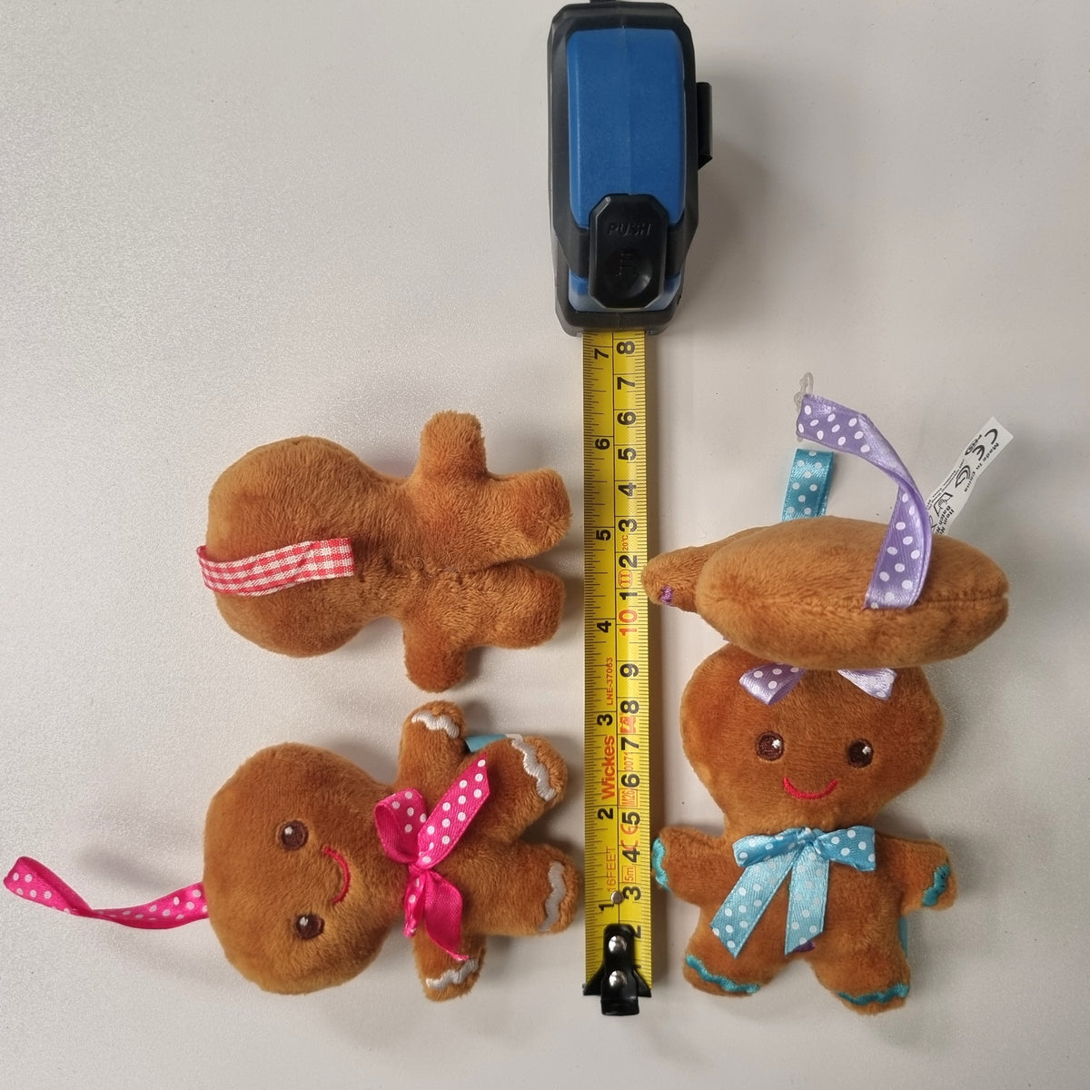 Three small brown plush gingerbread figures with colorful bows and hats are arranged on a light gray background with a yellow tape measure standing behind them for scale.