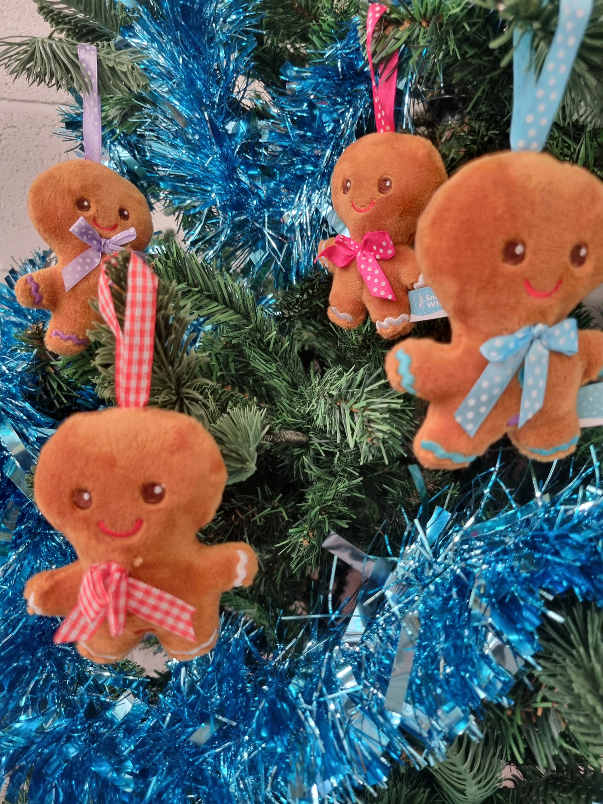 Four plush gingerbread man Christmas ornaments with embroidered faces and colorful ribbon bows hanging on a decorated Christmas tree with blue tinsel.