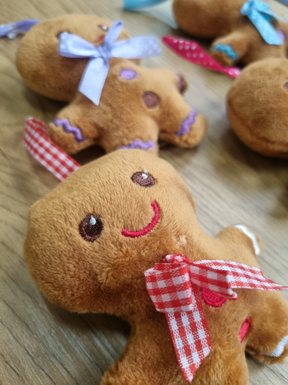 A close-up of a plush gingerbread person toy on a wooden surface, with other similar toys partially visible in the background. The main toy is brown with embroidered facial features, white trim, and a red-and-white gingham bow.