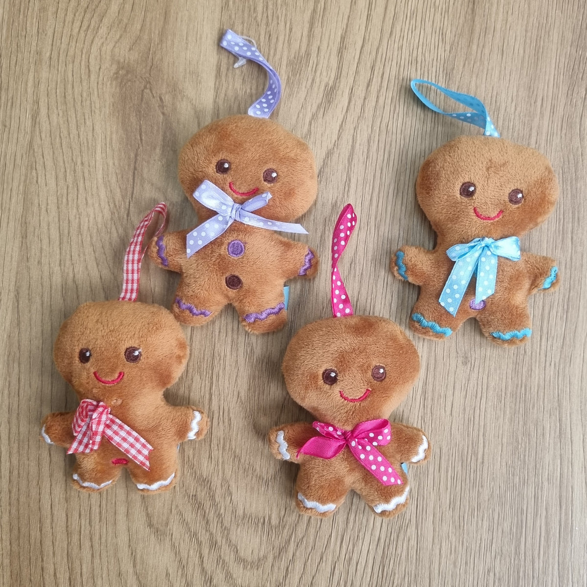 Four small gingerbread person plush ornaments in brown fabric, each with a colorful ribbon loop and bow, arranged on a light wood surface.