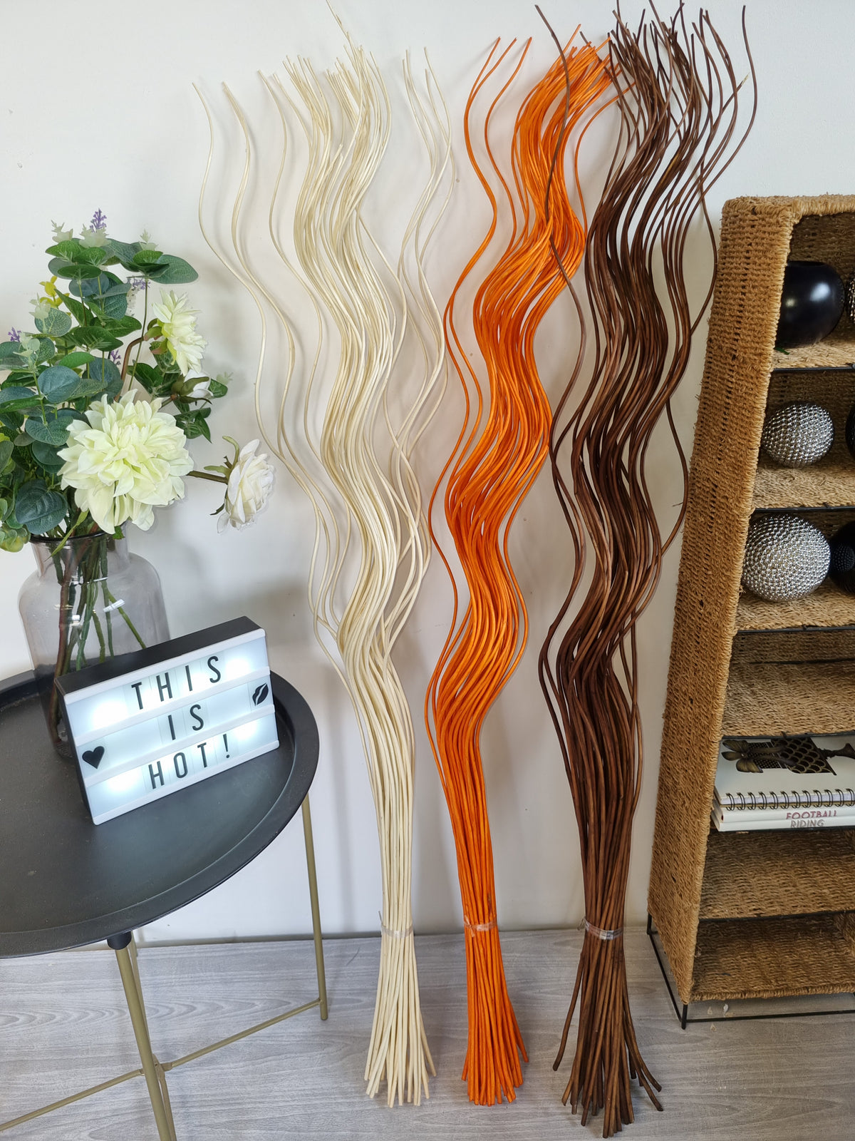 Three tall decorative wavy branches in cream, bright orange, and dark brown, displayed indoors next to a black side table with flowers and a woven shelf.