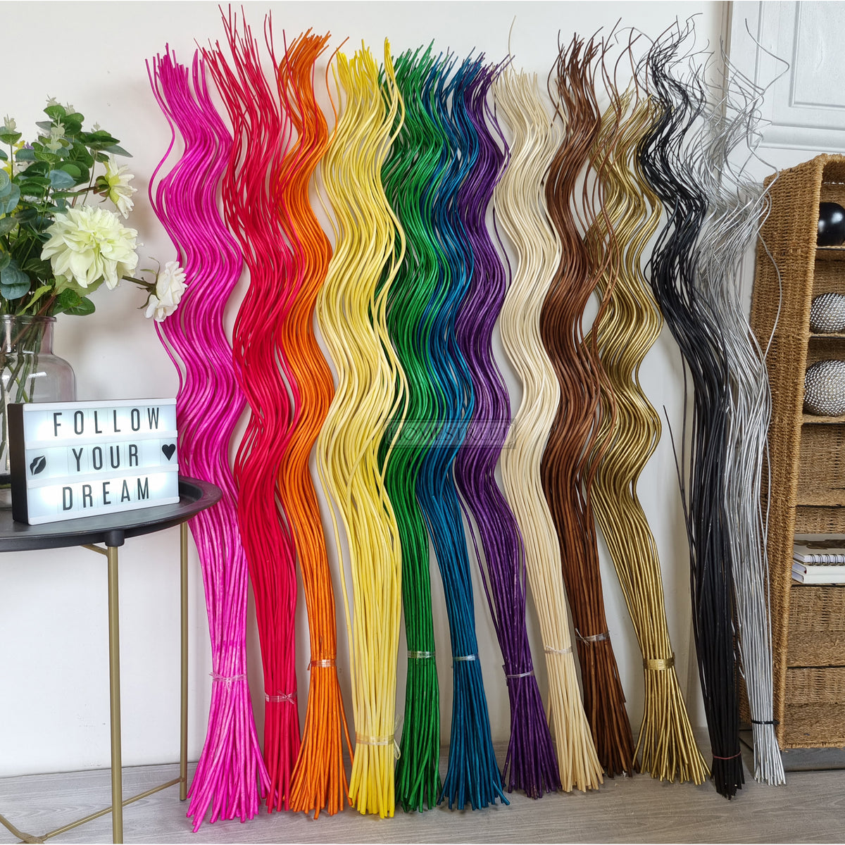 A selection of tall spiral willow twigs bunches dyed in various colors displayed vertically with a sign reading 'FOLLOW YOUR DREAMS' in the foreground.