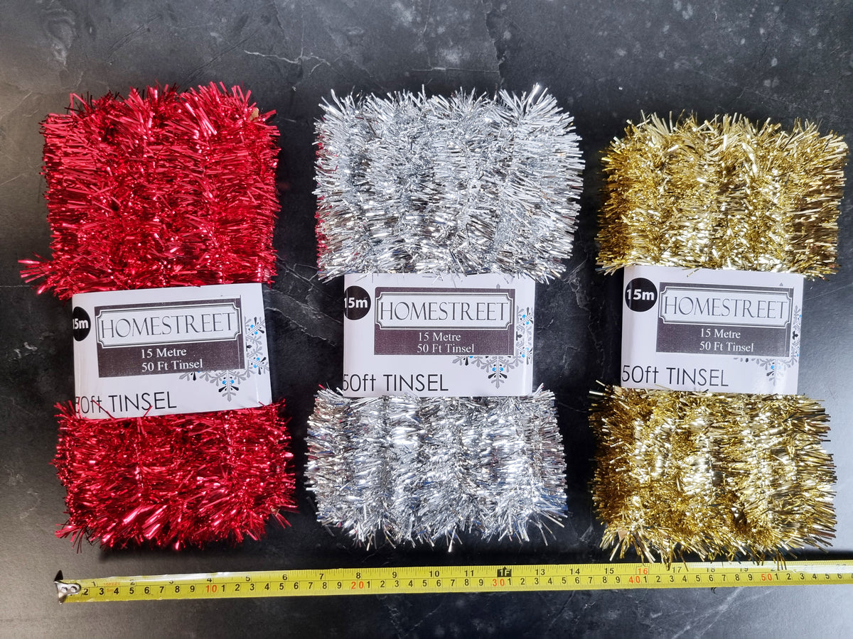 Three packages of Homestreet 50 foot Tinsel in red, silver, and gold, laid out side-by-side on a dark textured surface with a yellow measuring tape visible at the bottom.