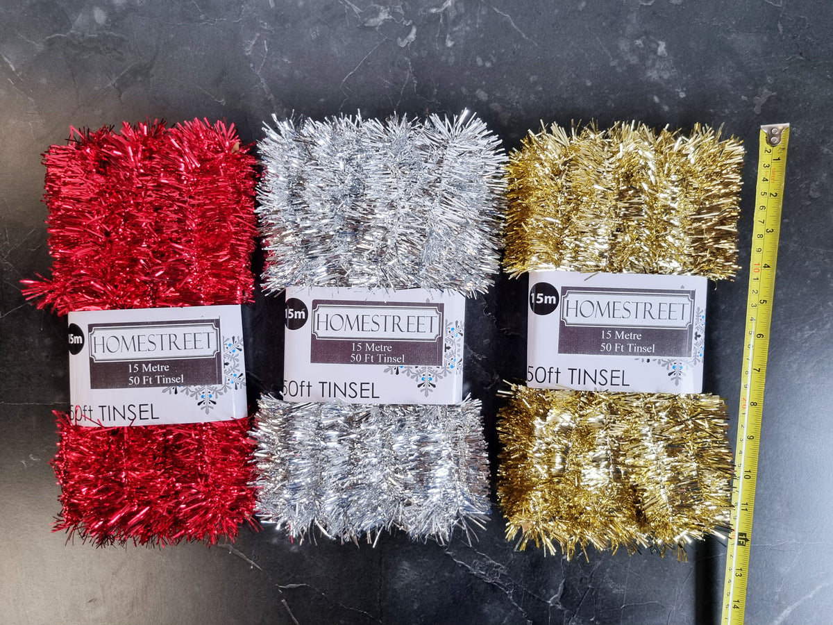 Three packages of Homstreet 50 foot Tinsel in red, silver, and gold, arranged side-by-side on a dark gray marble surface with a yellow measuring tape ruler on the right.