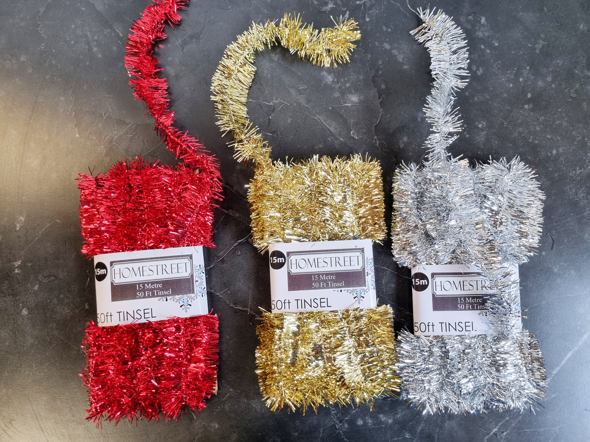 Three skeins of 15 metre tinsel yarn in red, gold, and silver, each with a white paper label reading 'HOME STREET' and '50ft TINSEL', arranged on a dark gray stone surface.