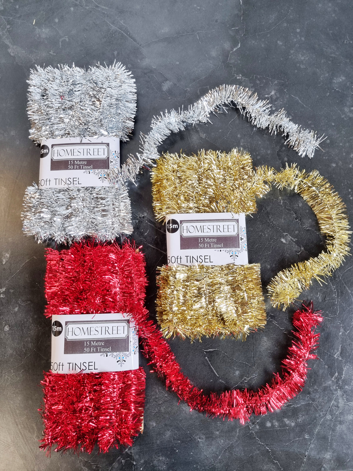 Three packages of Homestreet 15 foot long tinsel, one silver, one red, and one gold, displayed with strands coming out of the packs all on a dark gray stone surface.