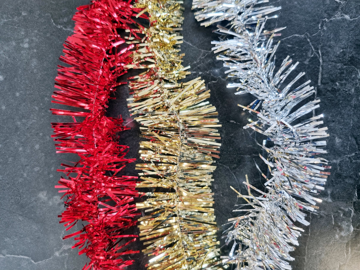 Three strands of metallic Christmas tinsel garland in red, gold, and silver, laid out side-by-side on a dark gray marble surface.