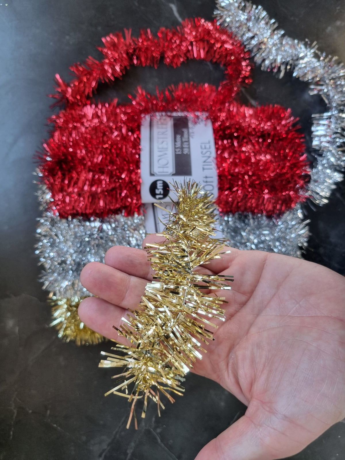 a hand holding gold tinsel to give an idea of the thickness with red and silver tinsel in the background on a black table
