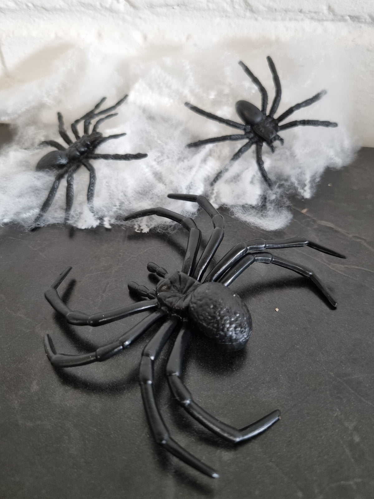 Three black plastic toy spiders arranged on a dark surface with white faux cobwebs in the background.
