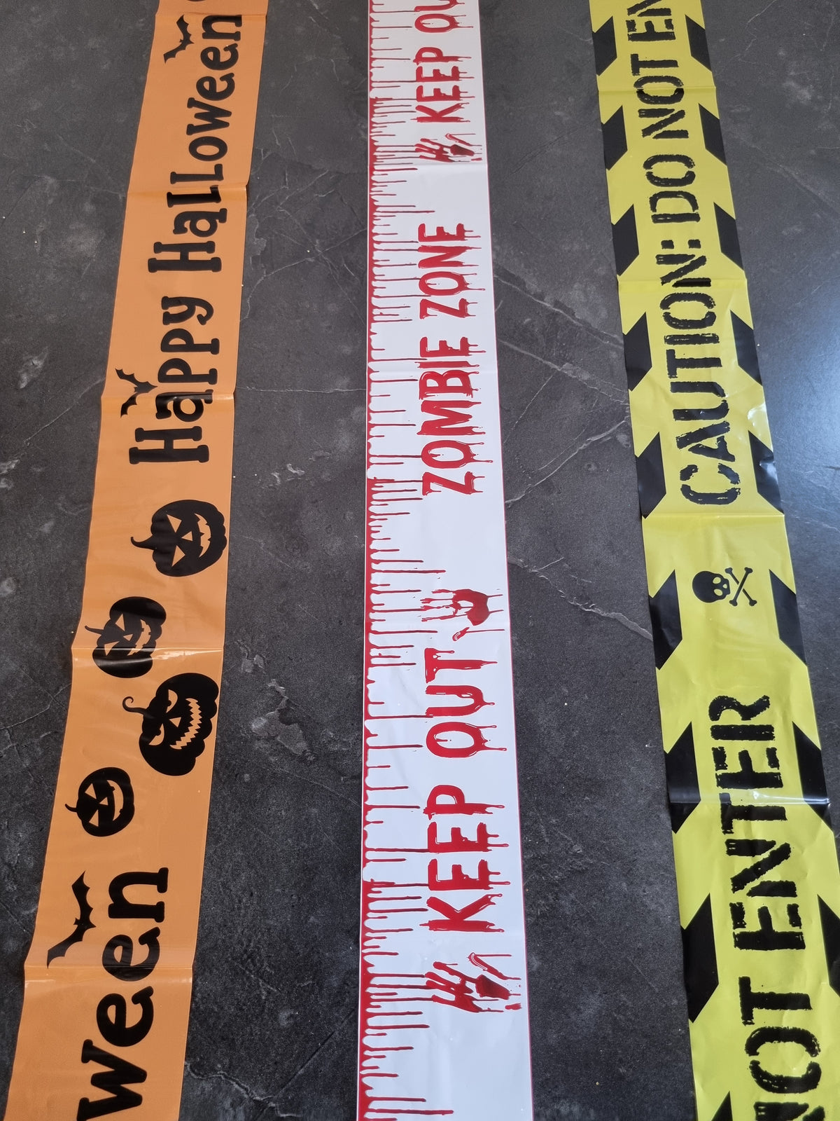 Three Halloween-themed decorative tape rolls laid out on a dark gray floor: orange tape with black 'Happy Halloween' text and jack-o'-lantern graphics, white tape with red 'KEEP OUT' and blood-spatter details, and yellow-and-black caution tape with 'CAUTION: DO NOT ENTER' text.