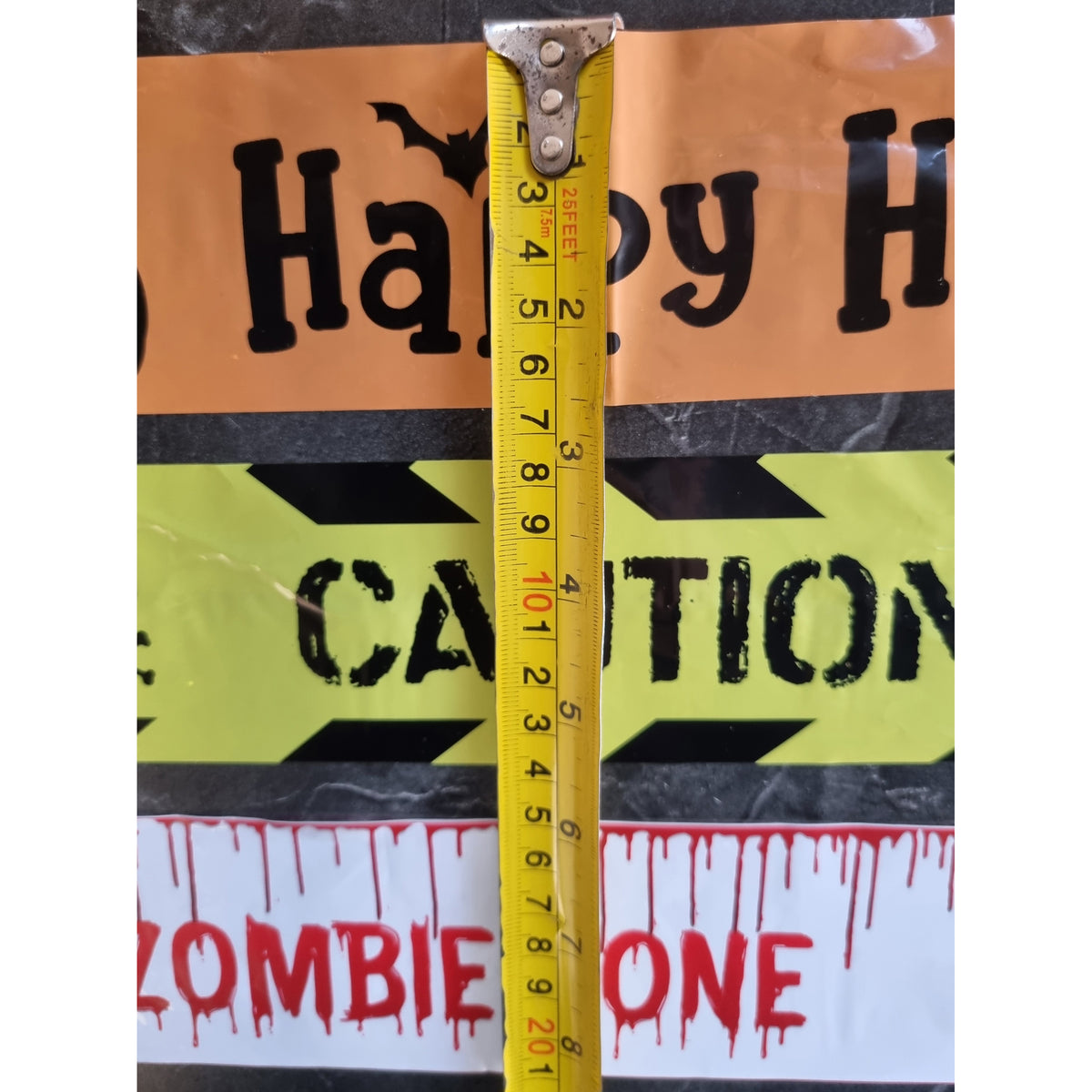 A yellow tape measure is shown vertically against Halloween warning signs that read 'Happy H' and 'CAUTION', with a white sign at the bottom reading 'ZOMBIE ONE'.