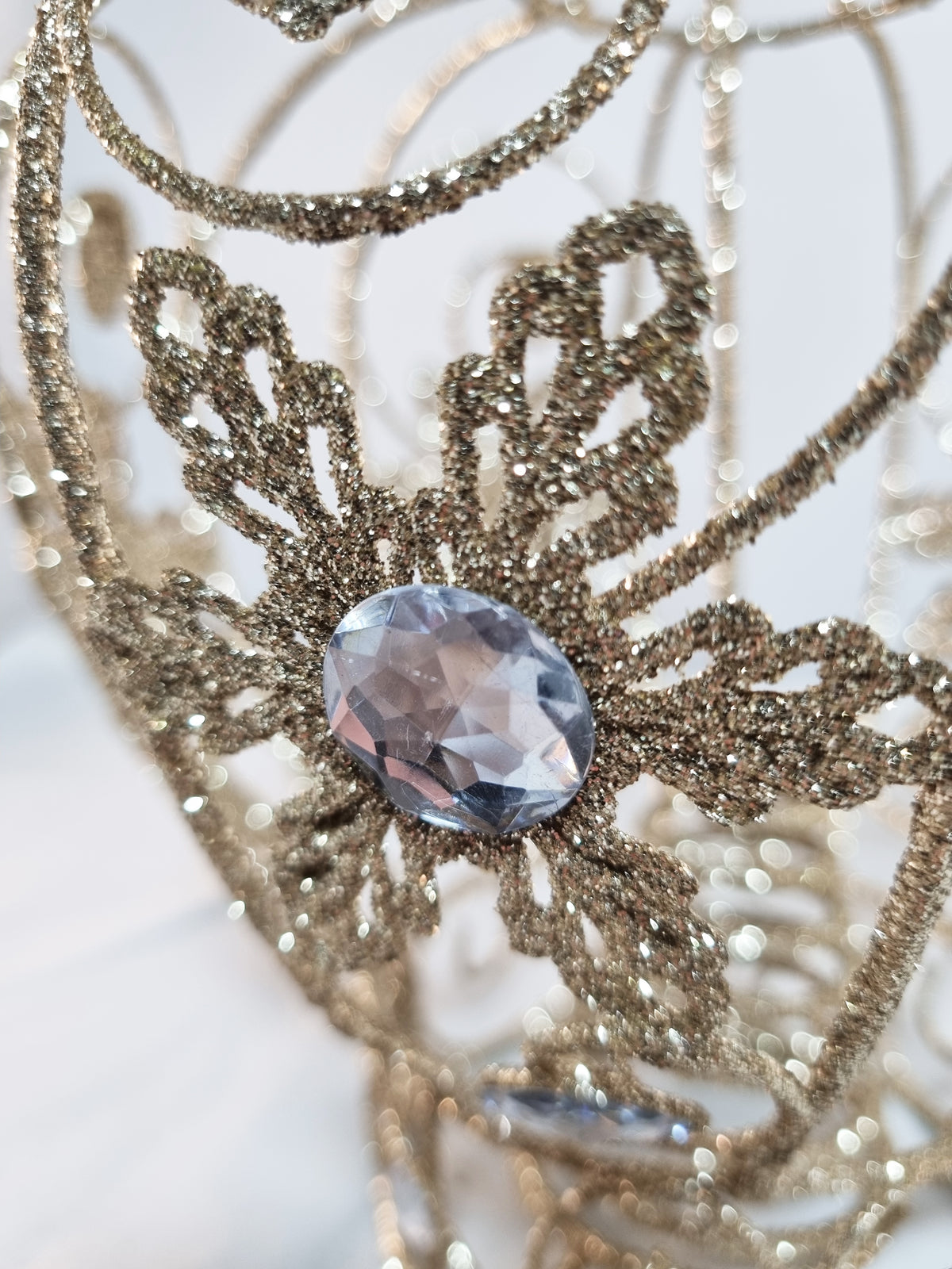 A close-up of a glittery gold fairy crown with a faceted light blue-gray crystal gemstone at the center, set against a soft white background.