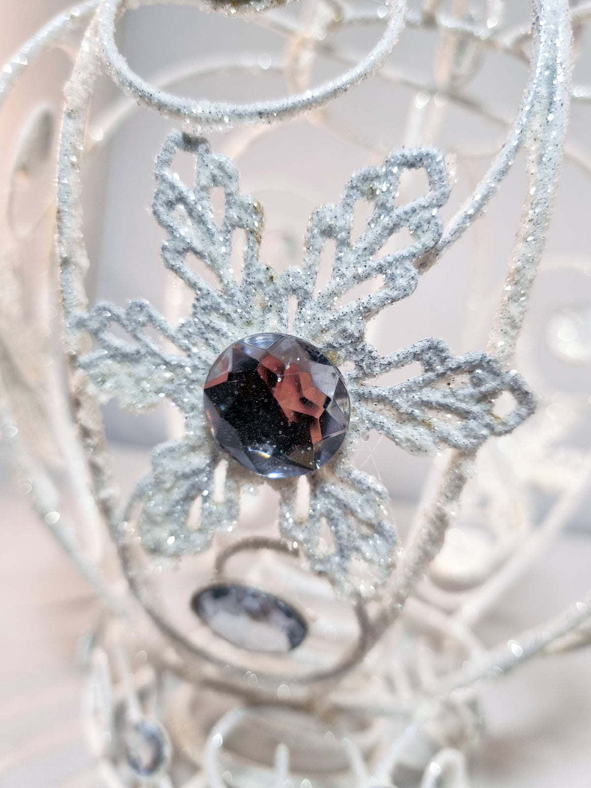 A close-up of a white metal glittery decorative crown tree topper or ornament featuring a central faceted clear acrylic gemstone and smaller matching stones, photographed against a soft light background.