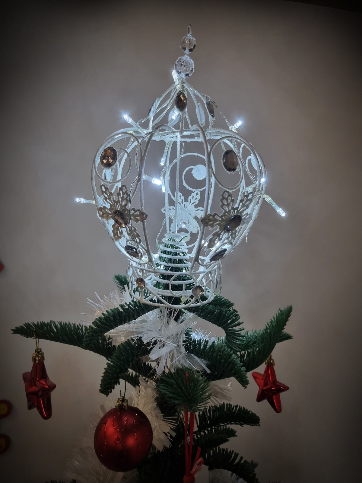 A white ornate Christmas tree topper with lit fairy lights and decorative swirls, displayed on top of a green artificial Christmas tree with red ornaments against a plain wall.