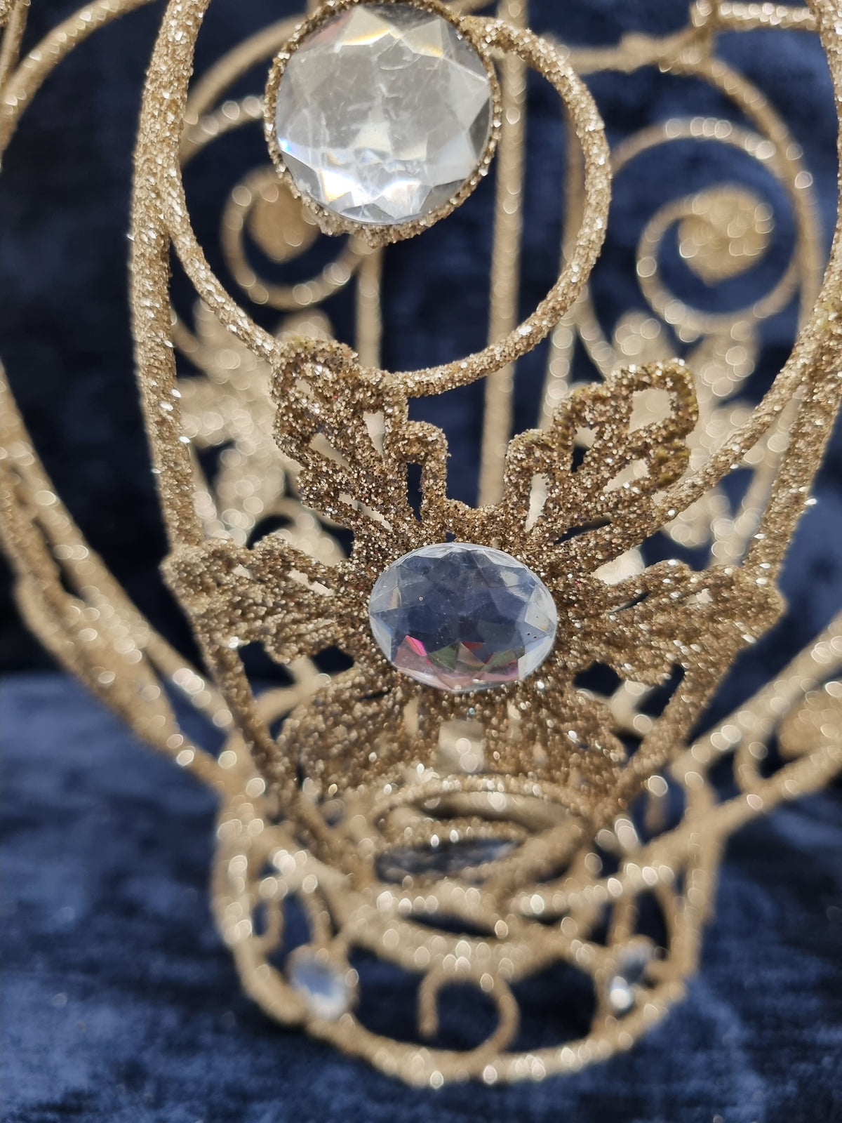 A close-up of an ornate gold glitter filigree headpiece or tiara with two faceted clear crystals, set against a dark blue velvet background.