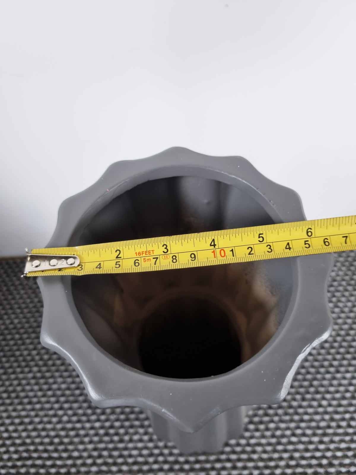A gray ceramic vase with a scalloped rim, shown from a top-down view with a yellow tape measure stretched across the opening to indicate its diameter.