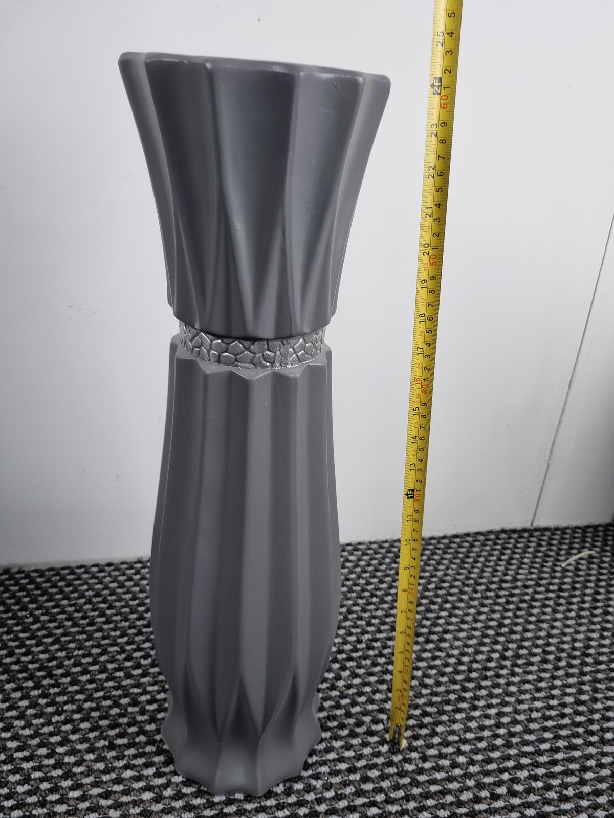 A gray ceramic vase with a flared top and textured vertical ridges, shown next to a yellow measuring tape on a black-and-white patterned rug against a white wall.