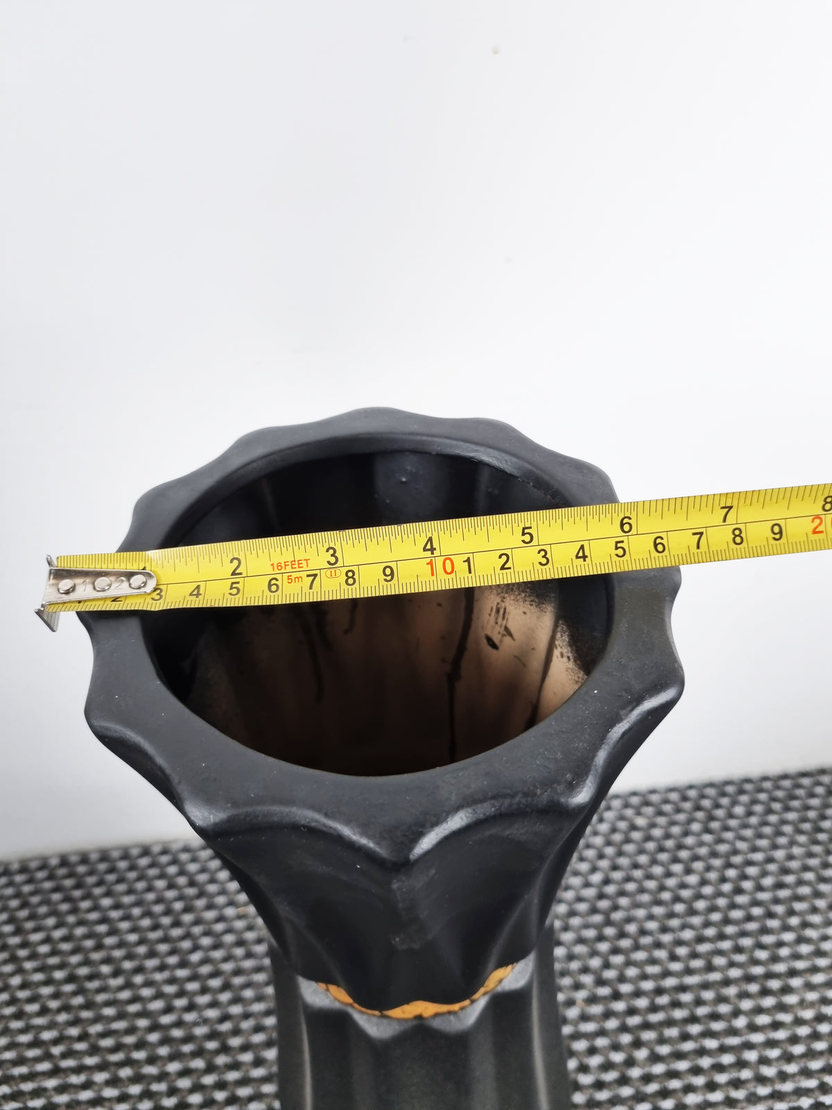 A black ceramic planter with a flared, textured rim, shown with a yellow tape measure stretched across the opening to indicate its width, set against a plain white background.