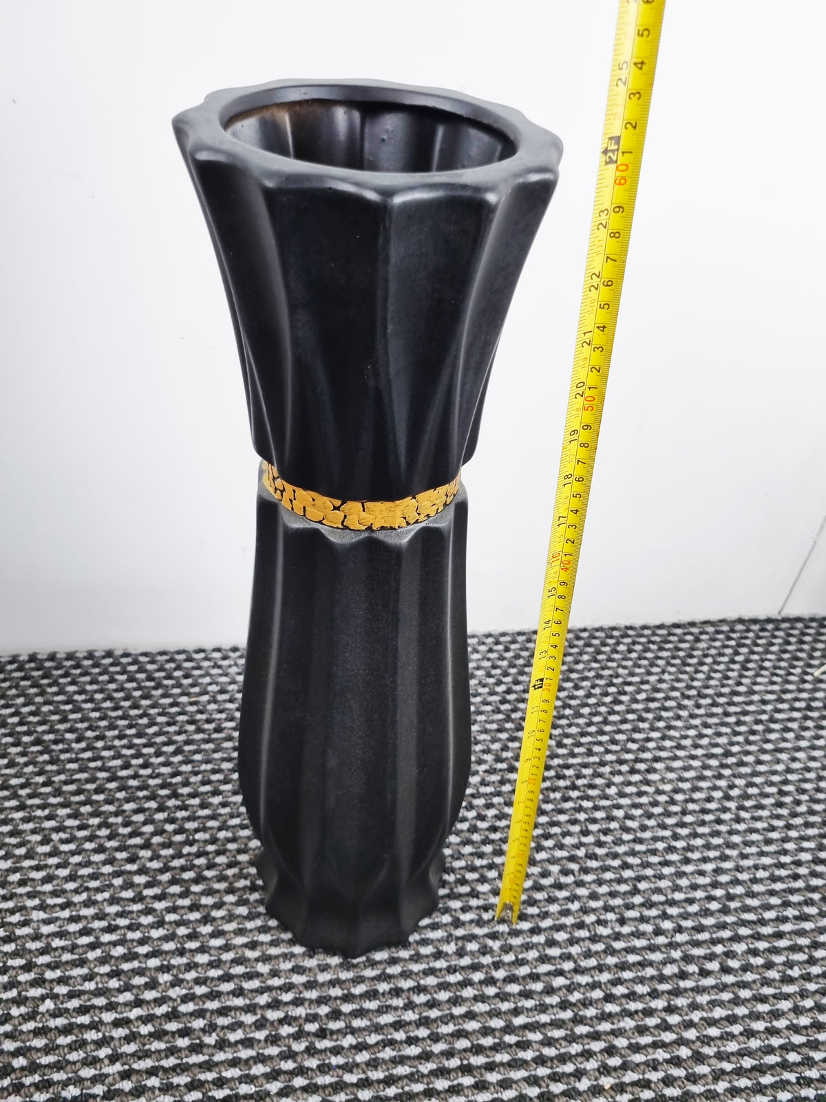 A black fluted ceramic vase with a textured finish, standing on a black and white patterned rug next to a yellow tape measure against a plain white wall.