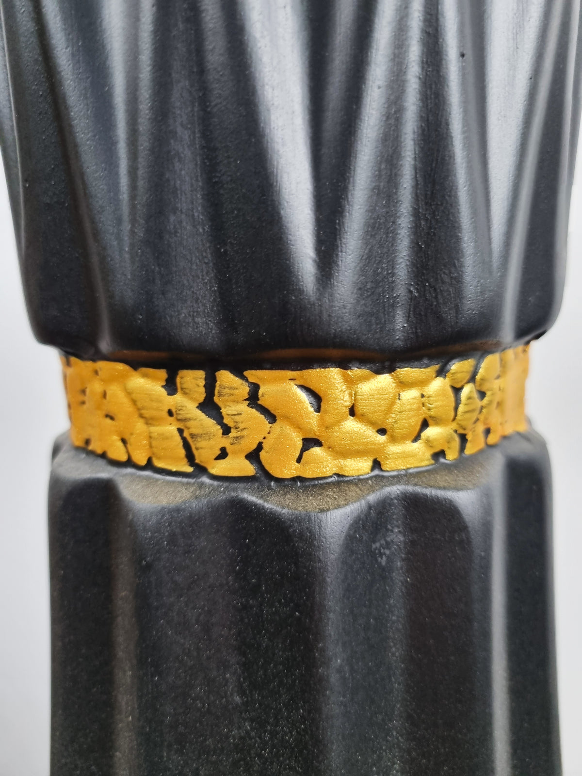 A close-up of a black ribbed decorative column or vase-like object with a wide textured gold band around the middle, shown against a light neutral background.