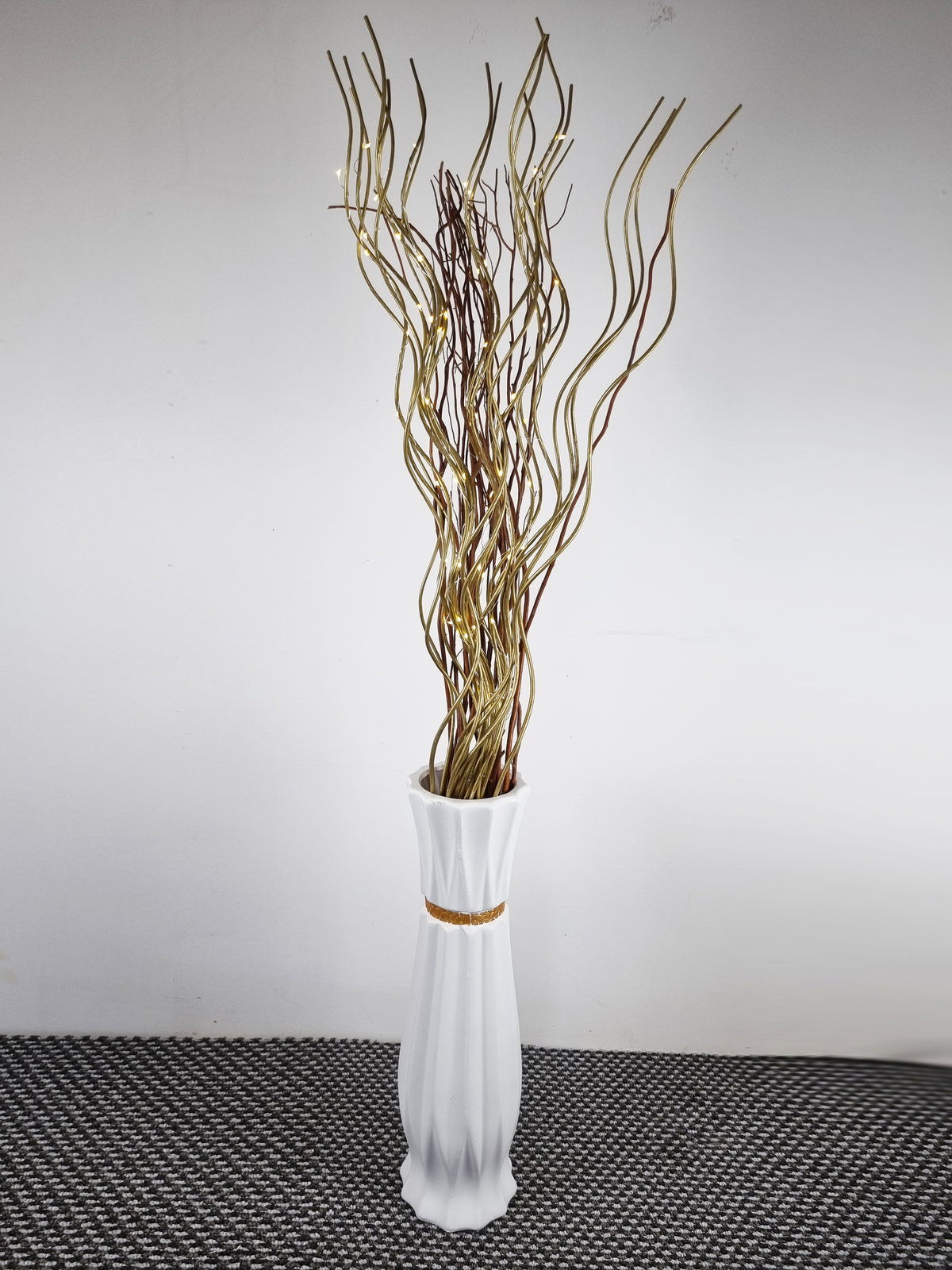 A tall white ceramic vase with a flared rim and a thin gold band around the middle, filled with dried branching branches in tan and brown tones, set against a plain light gray wall and dark textured floor.