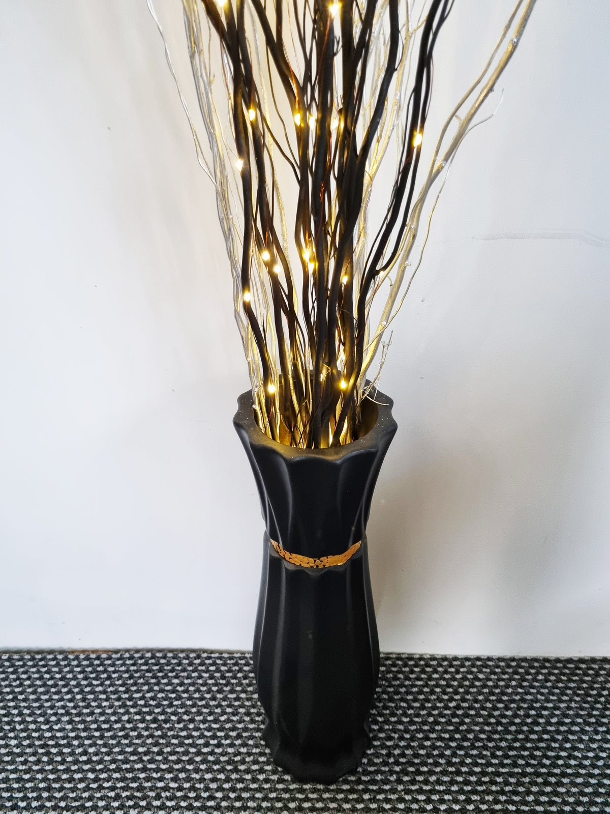 A black ceramic vase with a flared rim and a gold band detail, filled with decorative artificial branches and glowing warm white fairy lights, set against a plain white wall.