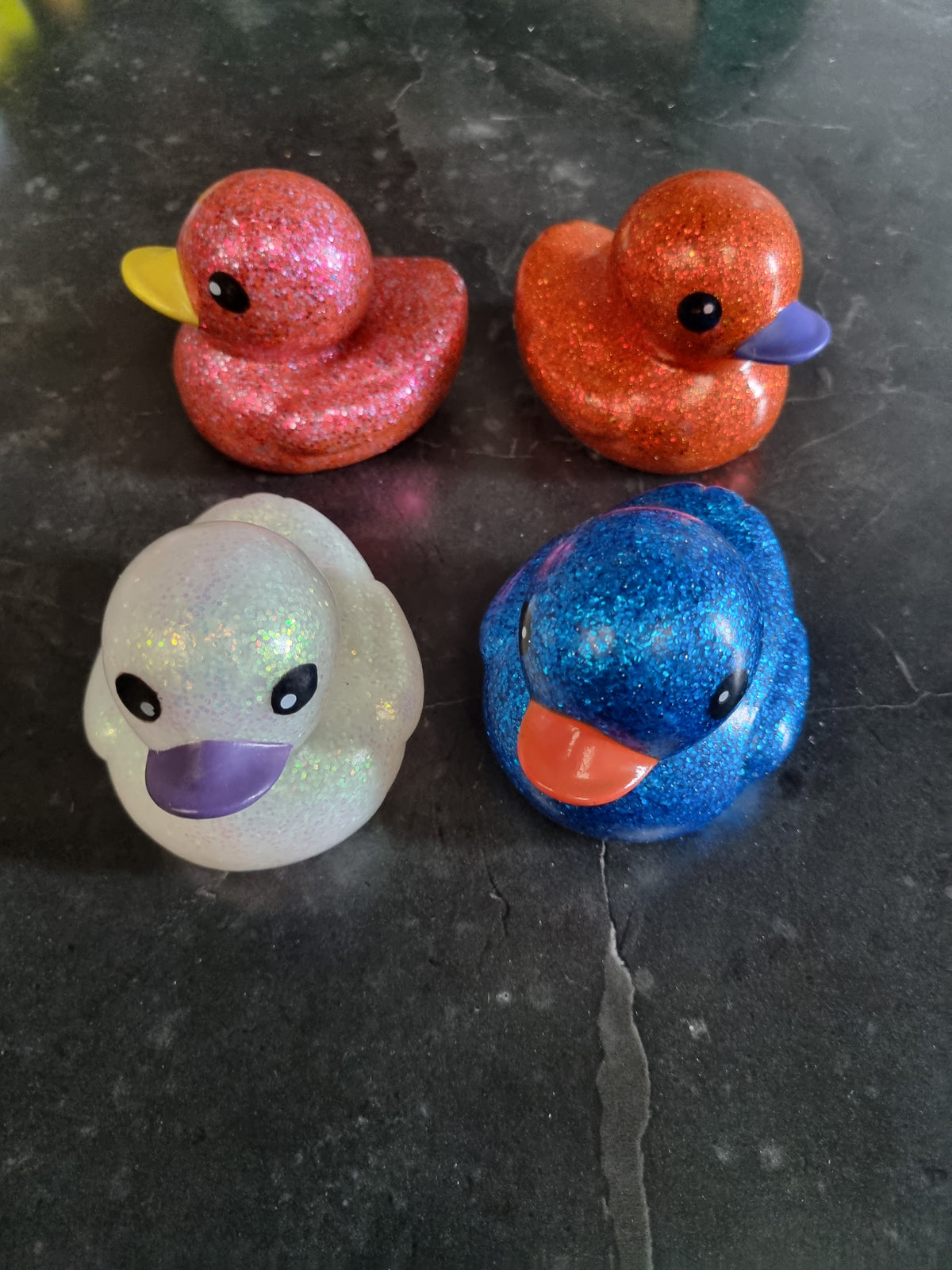 Four glittery rubber duckies arranged in a 2x2 grid on a dark gray stone surface. The top-left duck is pink glitter with a yellow beak, the top-right is orange glitter with a purple beak, the bottom-left is iridescent white with a purple beak, and the bottom-right is blue glitter with an orange beak.