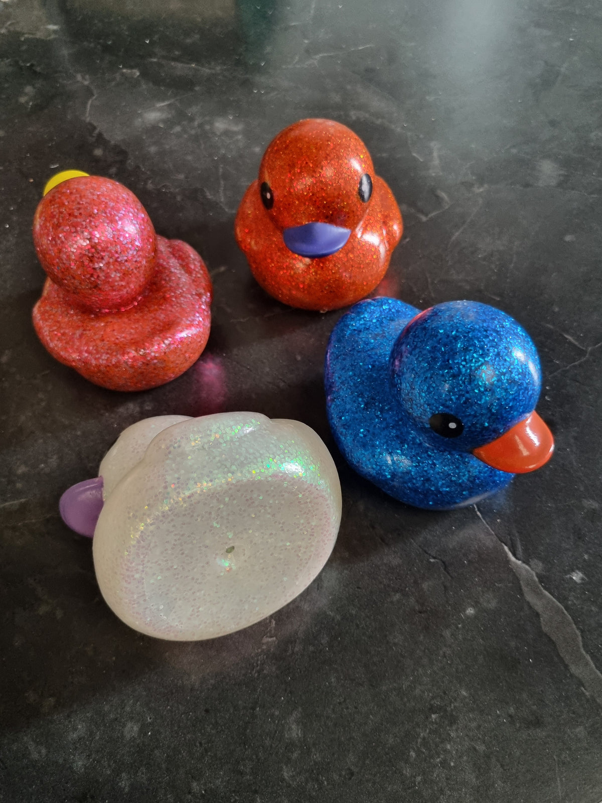 Four glittery rubber duck toys in red, orange, blue, and iridescent white, arranged on a dark gray stone surface.