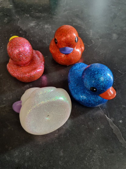 Four glittery rubber duck toys in red, orange, blue, and iridescent white, arranged on a dark gray stone surface.