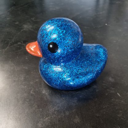 A small blue glitter rubber duck with a bright orange beak and a glossy black eye, sitting on a dark gray surface.
